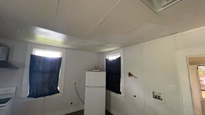 Una habitación con paredes blancas, una ventana con cortina azul, un refrigerador blanco y una puerta con un pequeño ventanal.