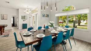 A dining area with a wooden table, blue chairs, and a chandelier. There is a window with a view of palm trees and a car outside. - 4BR Family Retreat with Pool, Lanai, Bikes & Games (Palm Coast)