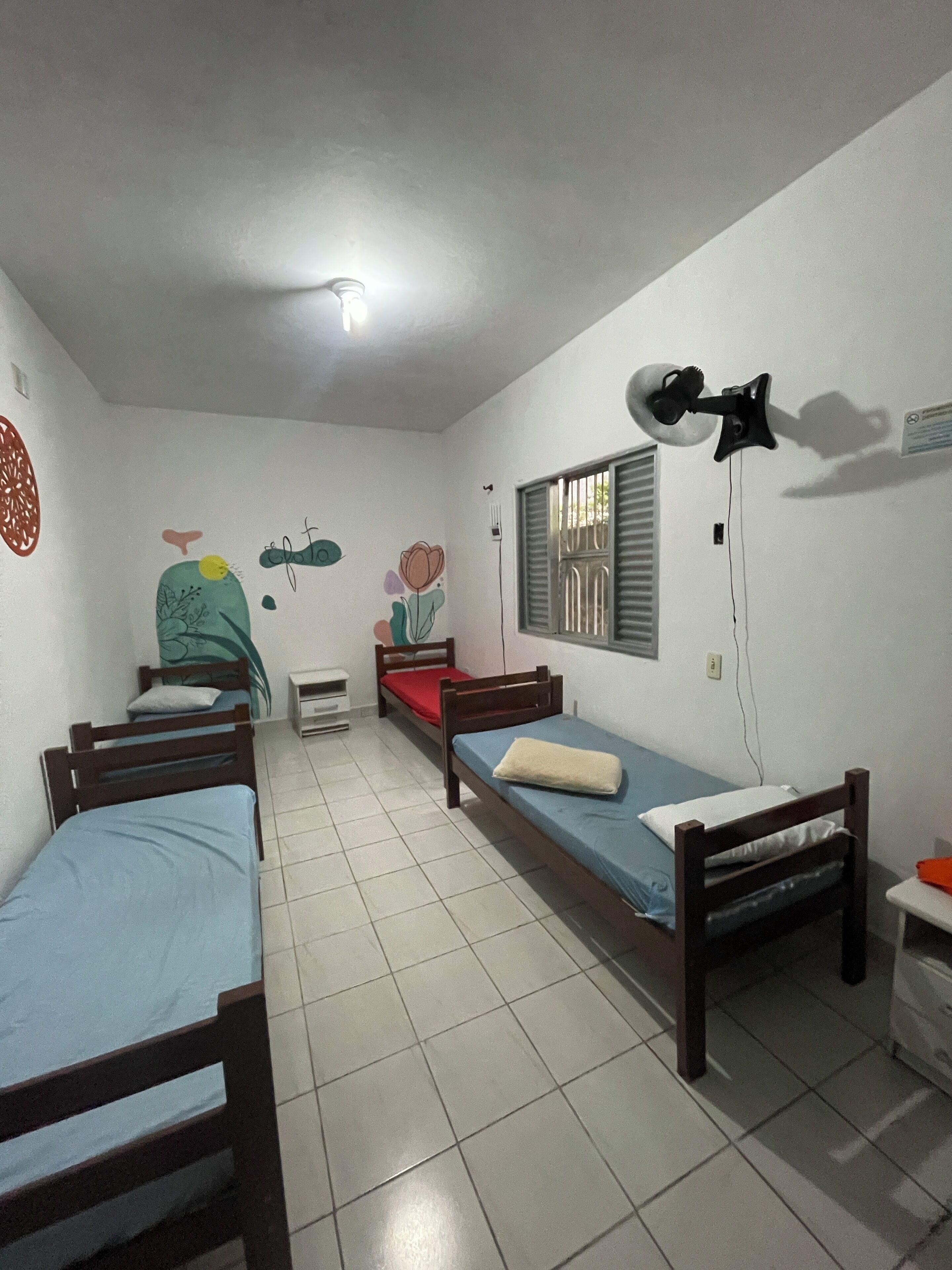 A dormitory room with three single beds, a ceiling fan, a window with bars, and a wall decoration.