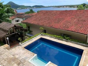 A swimming pool with a red-tiled roof, surrounded by greenery and a view of a river and mountains.