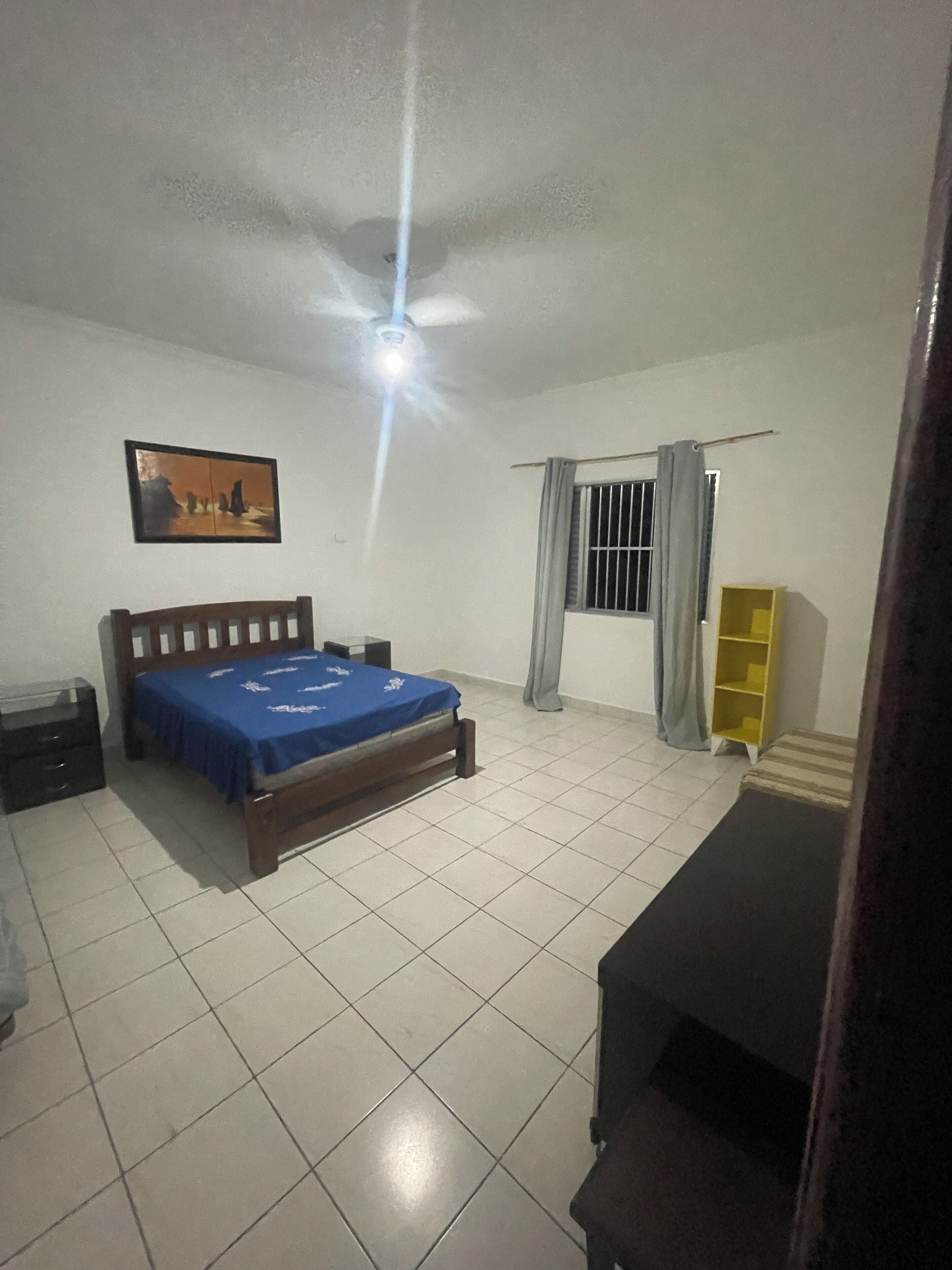Un dormitorio con una cama, una mesita de noche, un ventanal con cortinas, un ventilador de techo y un cuadro en la pared.