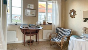 Ein gemütliches Zimmer mit Holzboden, einem runden Tisch mit Büchern und einer Vase, einem Korbstuhl mit gestreifter Sitzkissen und Blick auf ein rotdachiges Gebäude durch die Fenster.