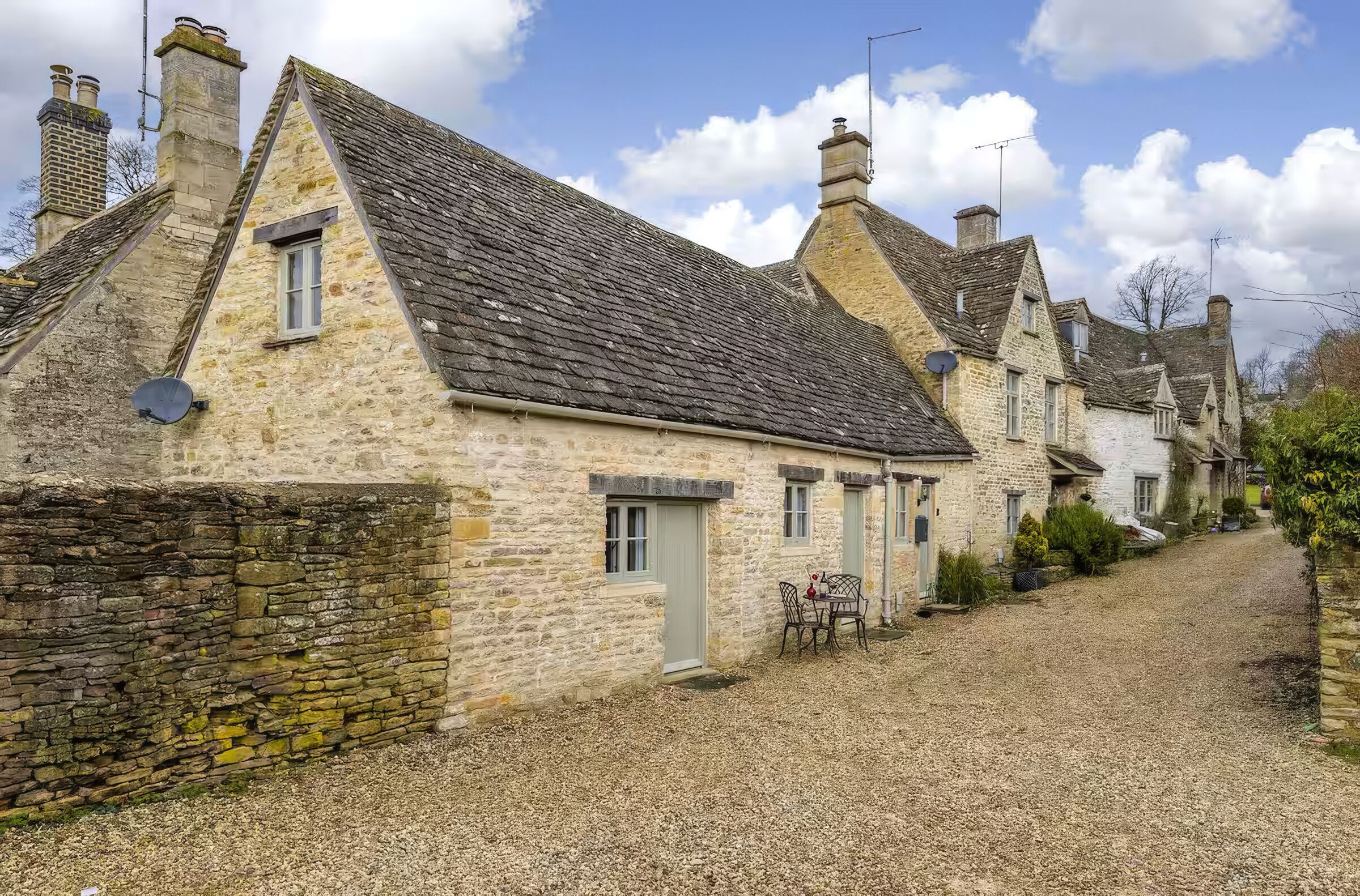 A row of stone houses with pitched roofs, a gravel driveway, and a small table with chairs outside one of the houses.