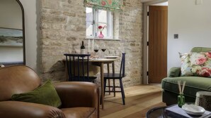 A cozy room with a stone wall, a dining table with chairs, a green sofa, and a window with a floral curtain. - Cosy Cotswold Cottage in Bibury w/ Private Parking - Perfect Romantic Getaway (Bibury)