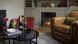 A cozy living room with a leather sofa, a glass coffee table with a bottle of champagne and glasses, a red ottoman, and a fireplace with a fire. - Cosy Cotswold Cottage in Bibury w/ Private Parking - Perfect Romantic Getaway (Bibury)