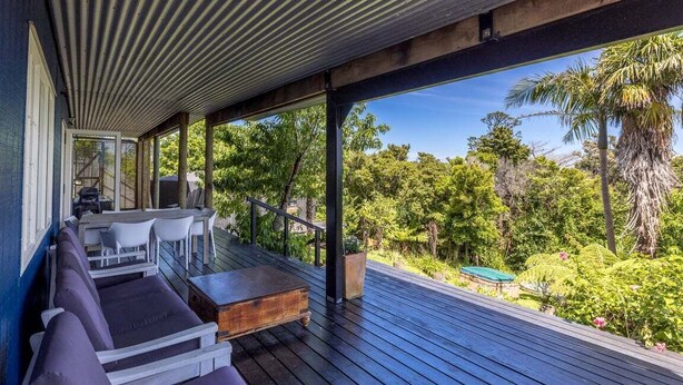 A covered outdoor seating area with wooden flooring, white and purple seating, and a view of a lush garden.