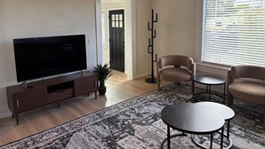 Un salon doté d’un téléviseur à écran plat, d’un meuble de rangement en bois foncé, d’un tapis à motifs, de deux fauteuils beiges, d’une petite table ronde et d’un grand lampadaire.