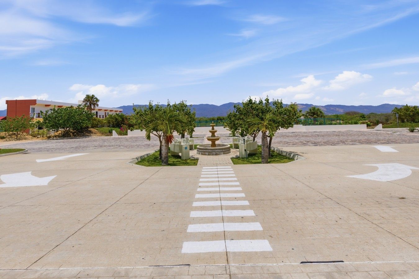 Un chemin pavé mène à une fontaine, bordé de chaque côté d’arbres, et s’étend dans un espace aménagé, avec des montagnes visibles au loin.