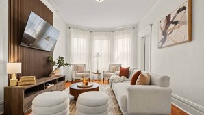 Ein modernes Wohnzimmer mit Flachbildfernseher, einer Couch, Hocker und einem Couchtisch.