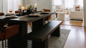 Ein Essbereich mit einem dunklen Holztisch, weißen Stühlen und einer Deckenleuchte. Auf dem Tisch stehen Kerzen und Gläser, und im Hintergrund eine weiße Couch.