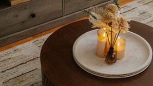 Ein runder Couchtisch aus Holz mit einer weißen Ablage, auf der Kerzen und getrocknete Blumen stehen. Im Hintergrund steht eine graue Holzkommode mit einer Pflanze und Büchern.