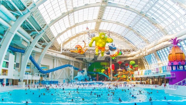 Ein großer, überdachter Wasserpark mit mehreren Schwimmbecken und Wasserrutschen, einer hohen Glasdecke und verschiedenen Wasserspielen.