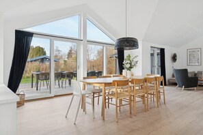 Un comedor amplio con una mesa grande y sillas, un candelabro colgante, y vistas al exterior a través de puertas de cristal.
