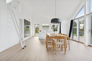 Una habitación amplia con una mesa de comedor, sillas, una lámpara colgante y vistas a través de amplias ventanas.
