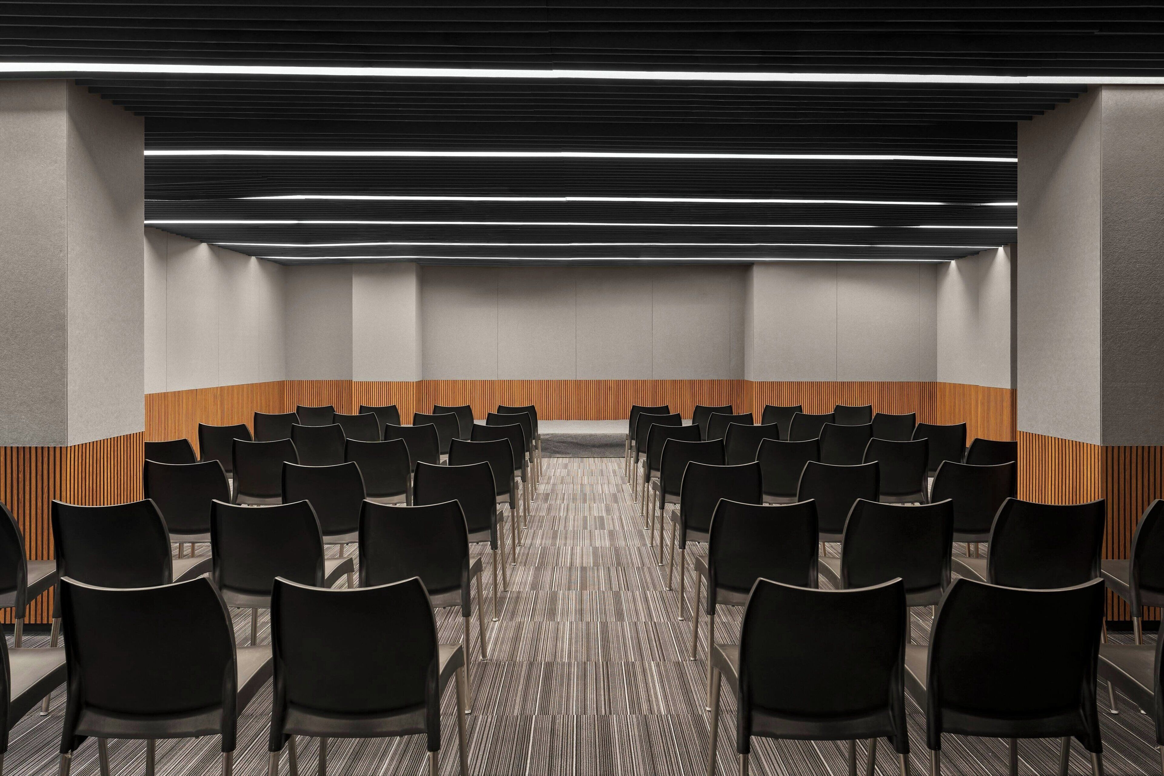 Un salón de conferencias con filas de sillas negras, piso alfombrado y una pared de acento de madera.