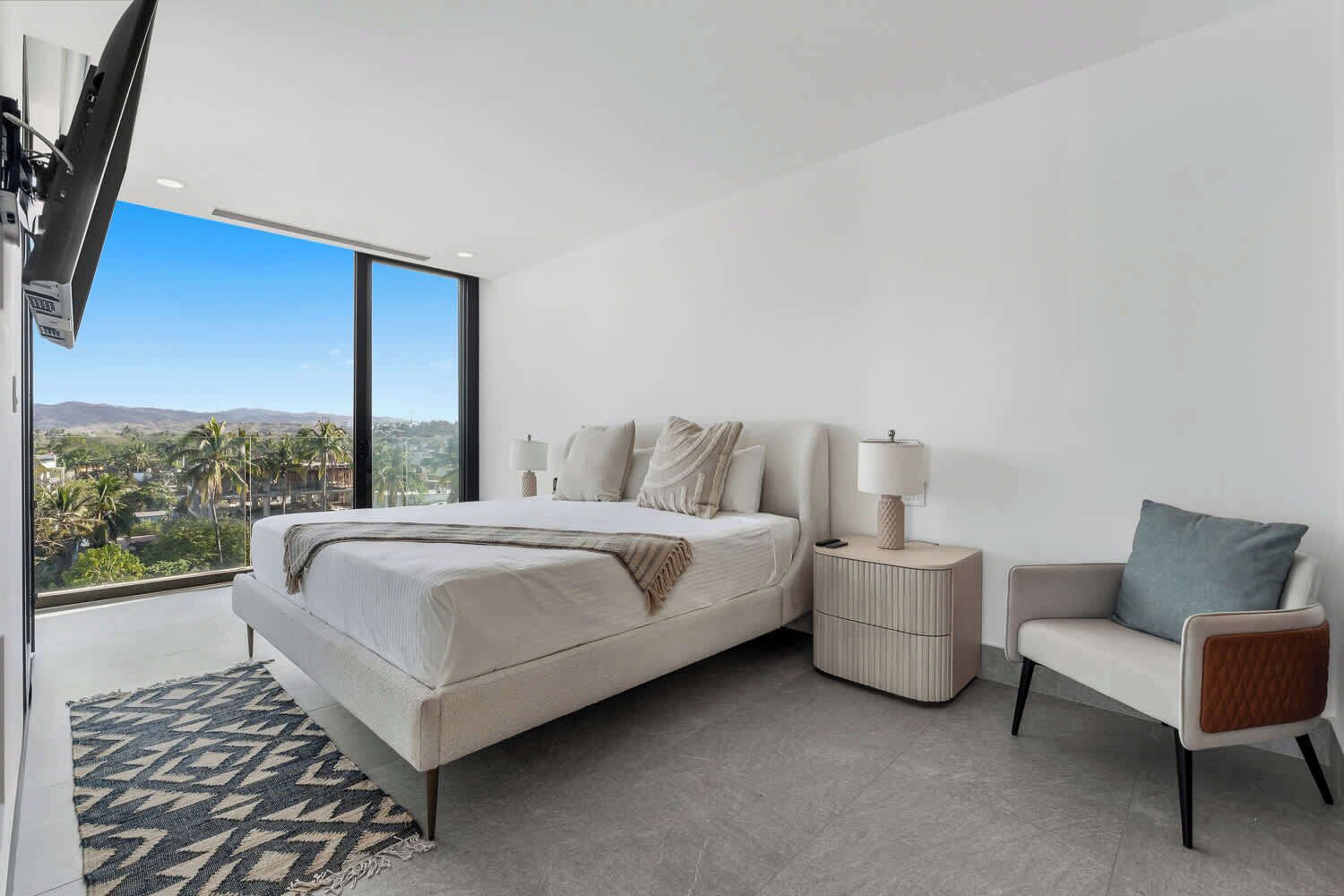 A modern bedroom with a large bed, bedside tables, a chair, and a view of palm trees and mountains.