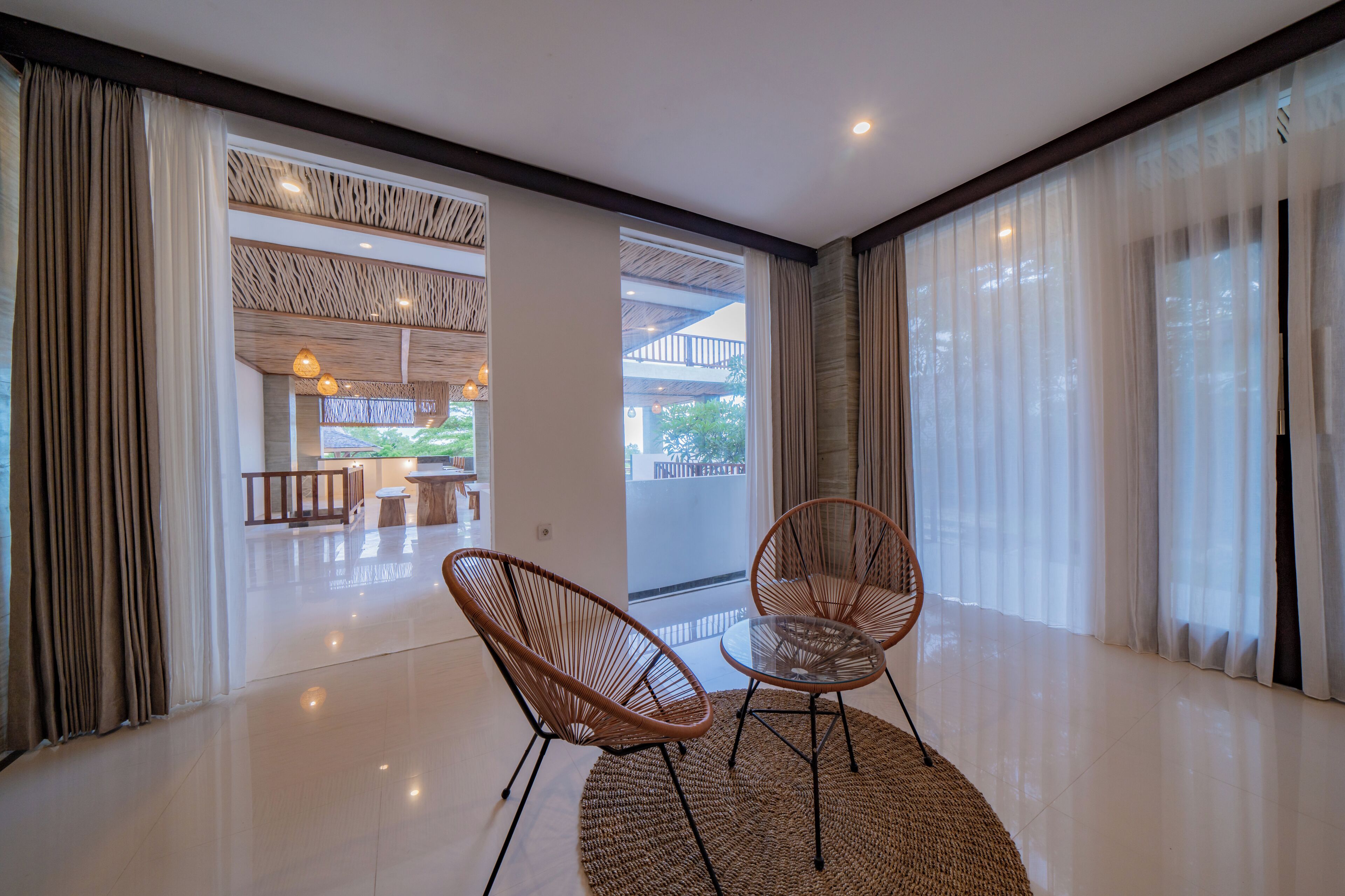 A spacious room with two wicker chairs and a glass table, large windows with sheer curtains, and a view of a balcony with outdoor furniture.