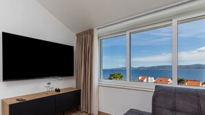 A modern living room with a large TV, a wooden console, a grey sofa, and a view of the sea and hills. - Apartman Irena by Interhome (Medici)