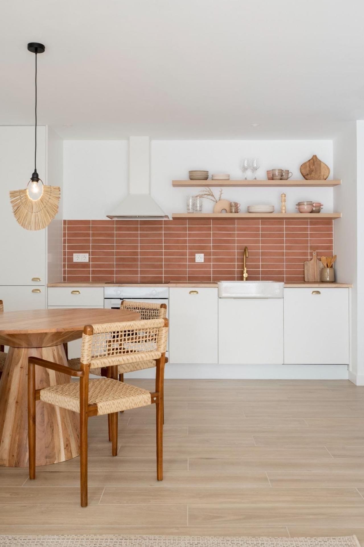 A modern kitchen with white cabinets, a wooden dining table, wicker chairs, and a hanging light fixture.
