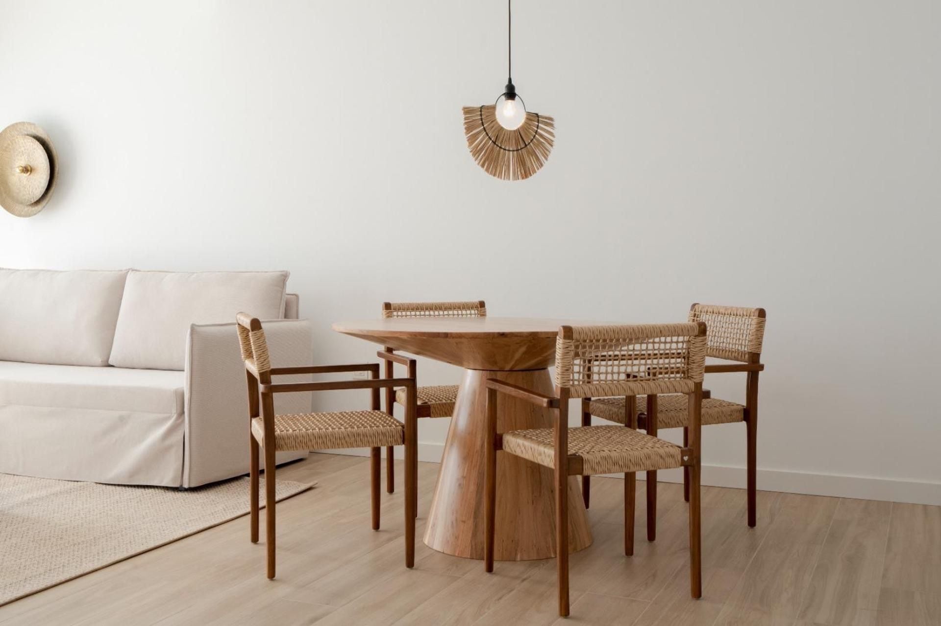 A minimalist living room with a round wooden table and four wicker chairs, a white sofa, and a hanging light fixture.