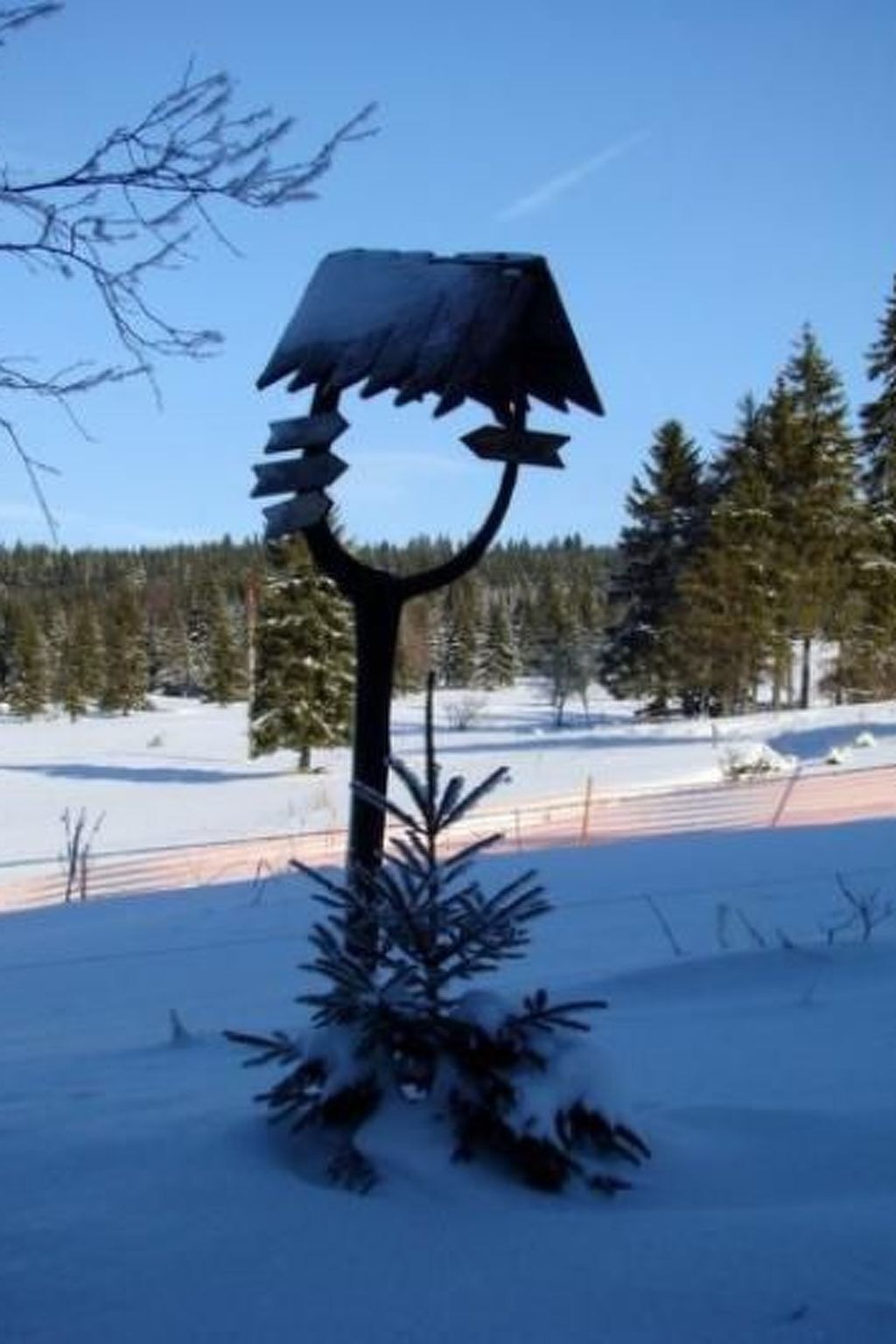 Una escultura de metal que imita un árbol con la parte superior cubierta de nieve, situada en un paisaje nevado con árboles perennes al fondo.