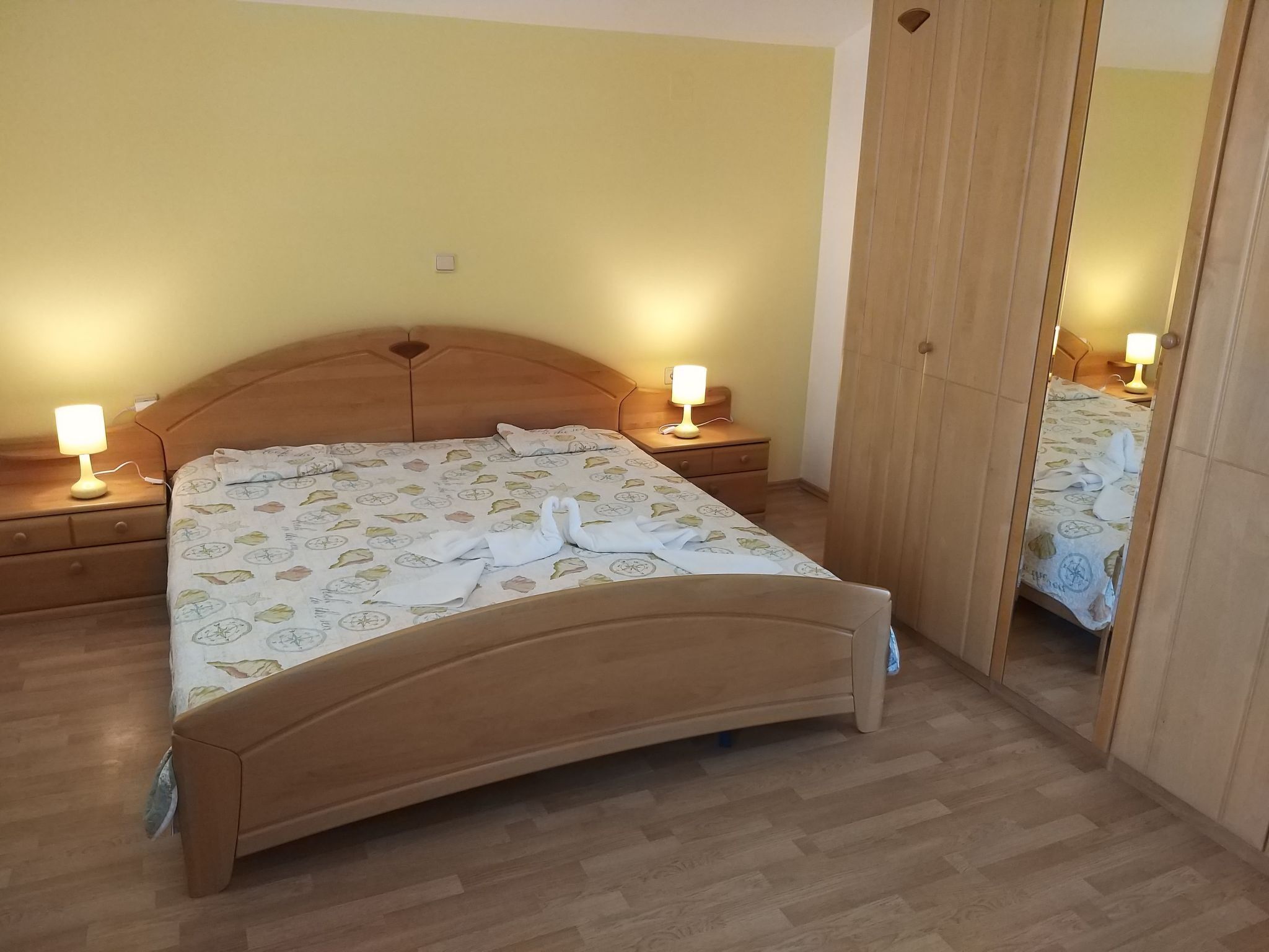 Ein ordentlich bezogenes Bett mit einem gemusterten Überwurf und zwei Handtüchern, die zu einer Schwanform arrangiert sind. Es gibt zwei Nachttische mit Lampen, einen Holzschrank und einen Spiegel.
