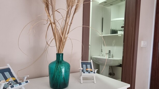 A bathroom vanity with a green vase and dried plants, a small white chair, and a mirror above a sink.