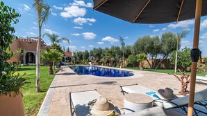 Ein Pool mit Liegestühlen und Sonnenschirmen, umgeben von Grünflächen und einem Gebäude im Hintergrund.