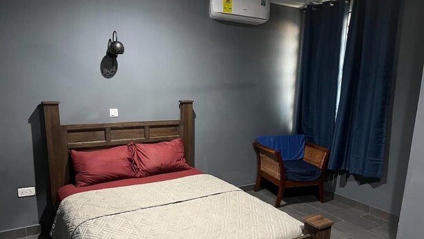 Een slaapkamer met een houten bed, rood-witte beddengoed, een wandairconditioner en een stoel met een blauwe hoes.