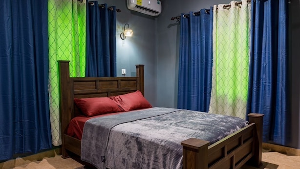 A bedroom with a wooden bed, red and grey bedding, blue curtains, and a wall-mounted air conditioner. - Clean beds, Sound Sleep (Accra)