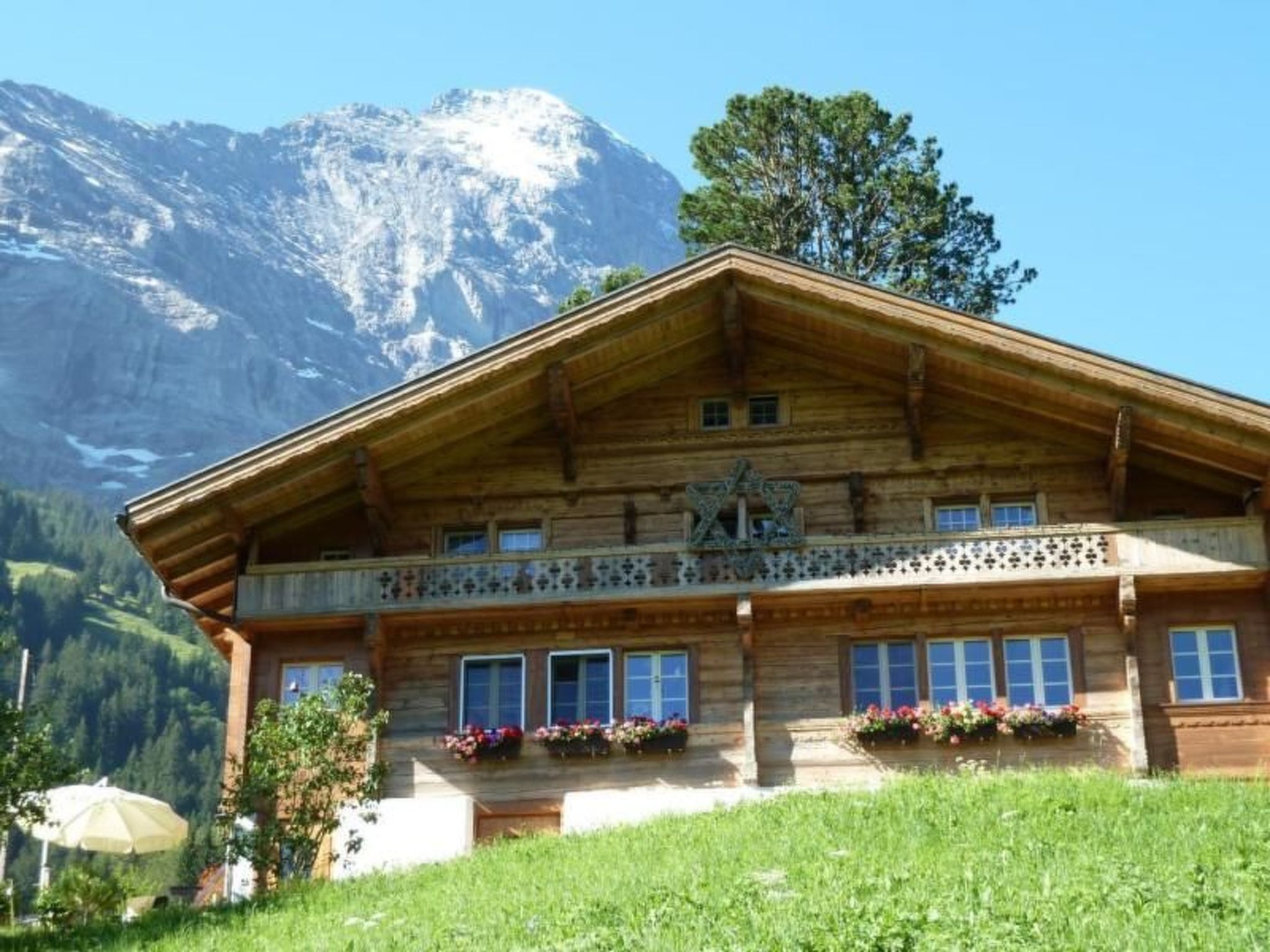 A wooden chalet with a balcony, windows, and flower boxes, set against a mountainous backdrop.