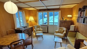 A cozy living room with wooden walls, a dining table, two armchairs, a sofa, and a bookshelf.