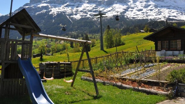 Een speeltuin met een glijbaan en schommels, een houten constructie, en een uitzicht op besneeuwde bergen op de achtergrond.
