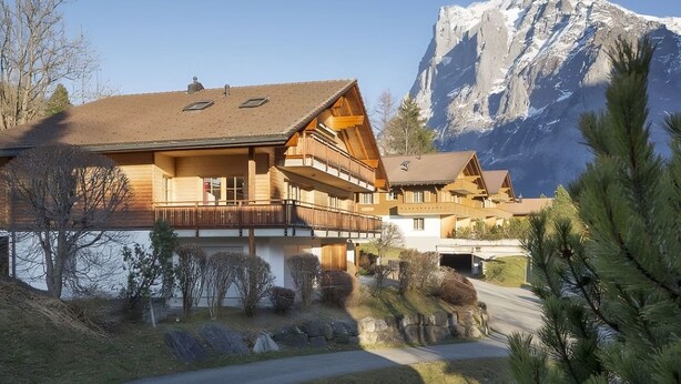 Un edificio de estilo chalete con fachada de madera y varios balcones, ubicado junto a un lago con montañas al fondo.