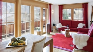 Amplia sala de estar con un sofá rojo, una mesa de comedor de madera con frutas y ventanas grandes con cortinas rojas.