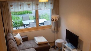 Ein modernes Wohnzimmer mit einem Ecksofa, einem Fernseher auf einem Schrank, einer Stehlampe und einem Fenster mit Vorhängen.