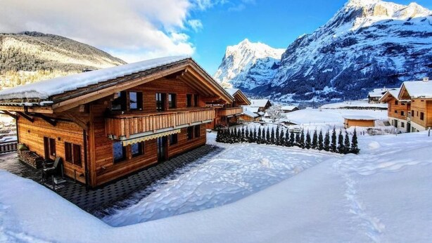 A wooden chalet with a balcony, surrounded by snow-covered landscape and mountains.