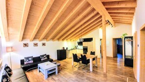 A spacious living area with a high wooden ceiling, a dining table with chairs, a black sofa, and a small coffee table.