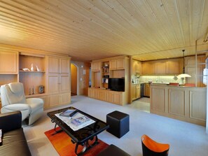 En rummelig opholdsstue med sofa, sofabord og fjernsyn. Der er et køkkenområde med skabe og et spisebord.