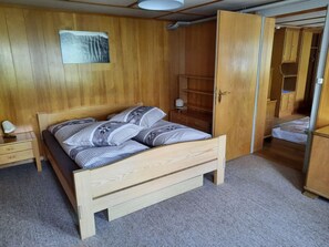 Een houten slaapkamer met een bed, nachtkastje, bedlamp, kledingkast en wandplank.