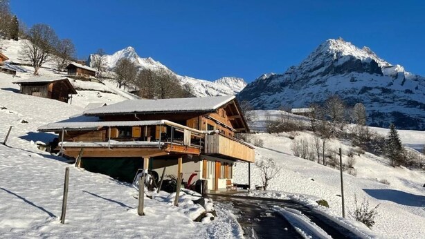 A wooden chalet with a balcony, set against a backdrop of snow-covered mountains and a clear blue sky. - Chalet Bachsbort 14 by Interhome (Grindelwald)