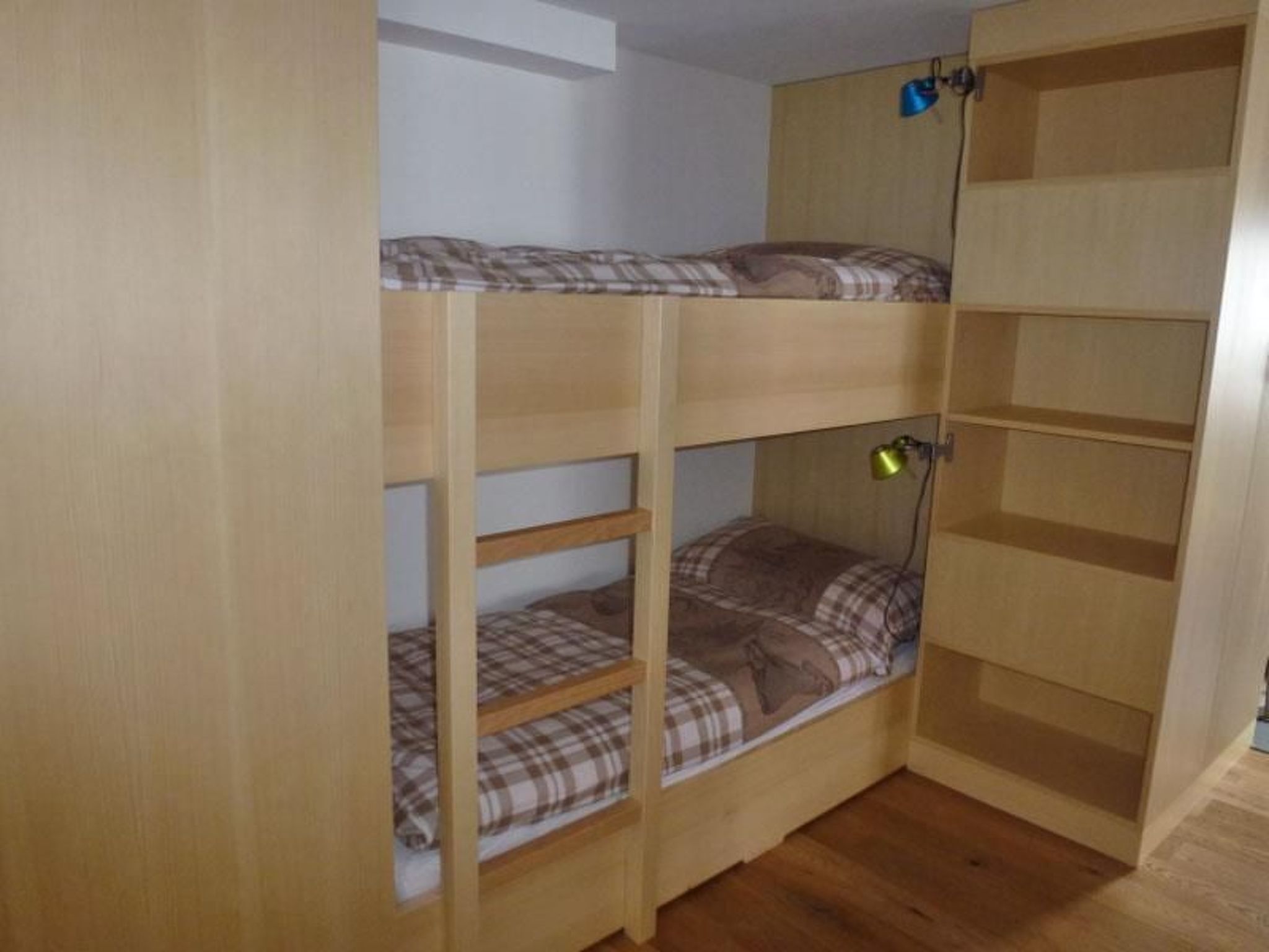 Een stapelbed met een matras met ruitdessin, een klein bureau met lamp en een houten garderobe.