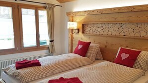 Ein Doppelzimmer mit zwei Betten, jeweils mit karierten Bettbezügen und roten Kissen. Es gibt ein Fenster mit Vorhängen, eine Wandlampe und einen Kopfteil mit Steinmuster.