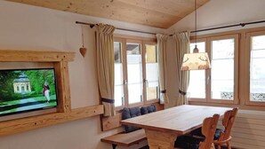 Ein gemütliches Zimmer mit einem Holzesstisch, einer Sitzbank, einem an der Wand montierten Fernseher und großen Fenstern mit Vorhängen.