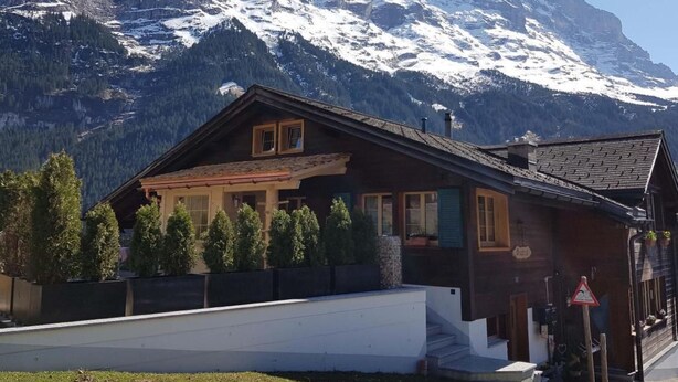 Eine Holzhütte mit geneigtem Dach, einer Veranda mit Geländer und Blick auf verschneite Berge im Hintergrund.