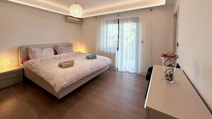 Ein Schlafzimmer mit einem großen Bett, Nachttischlampen, einer Deckenleuchte, einem Fenster mit durchsichtigen Vorhängen und einem kleinen Tisch mit einer Vase.