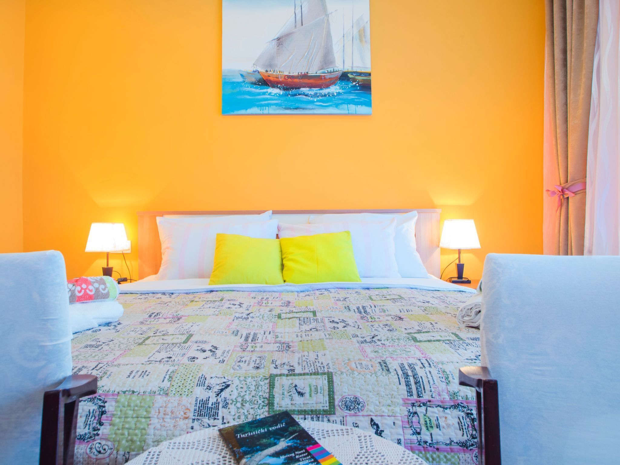 Une chambre d’hôtel avec un mur orange, un lit recouvert d’une courtepointe colorée, deux lampes de chevet et un tableau représentant un voilier.