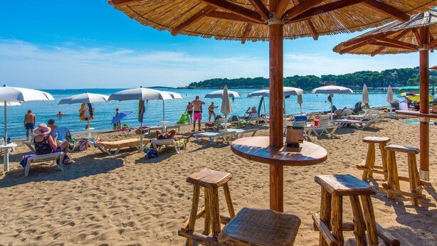 Ein Strand mit Holztischen und -stühlen, Sonnenschirmen und entspannenden Menschen.