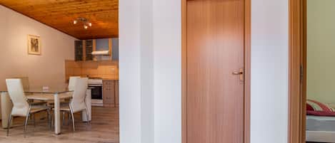 A modern interior with a wooden ceiling, a dining area with a glass table and chairs, a kitchen with a stove and cabinets, and a wooden door.