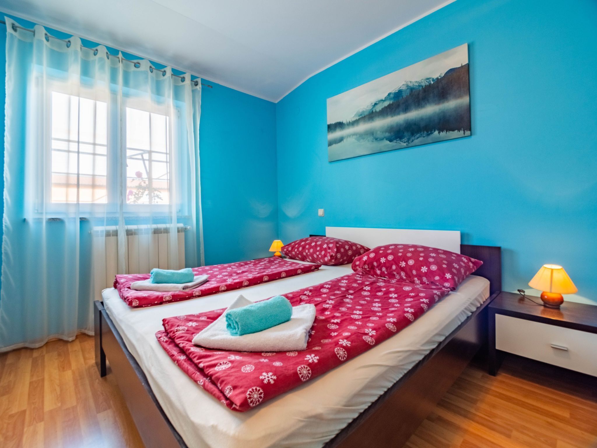 A bedroom with a large bed, two bedside lamps, a window with sheer curtains, and a painting on the wall.